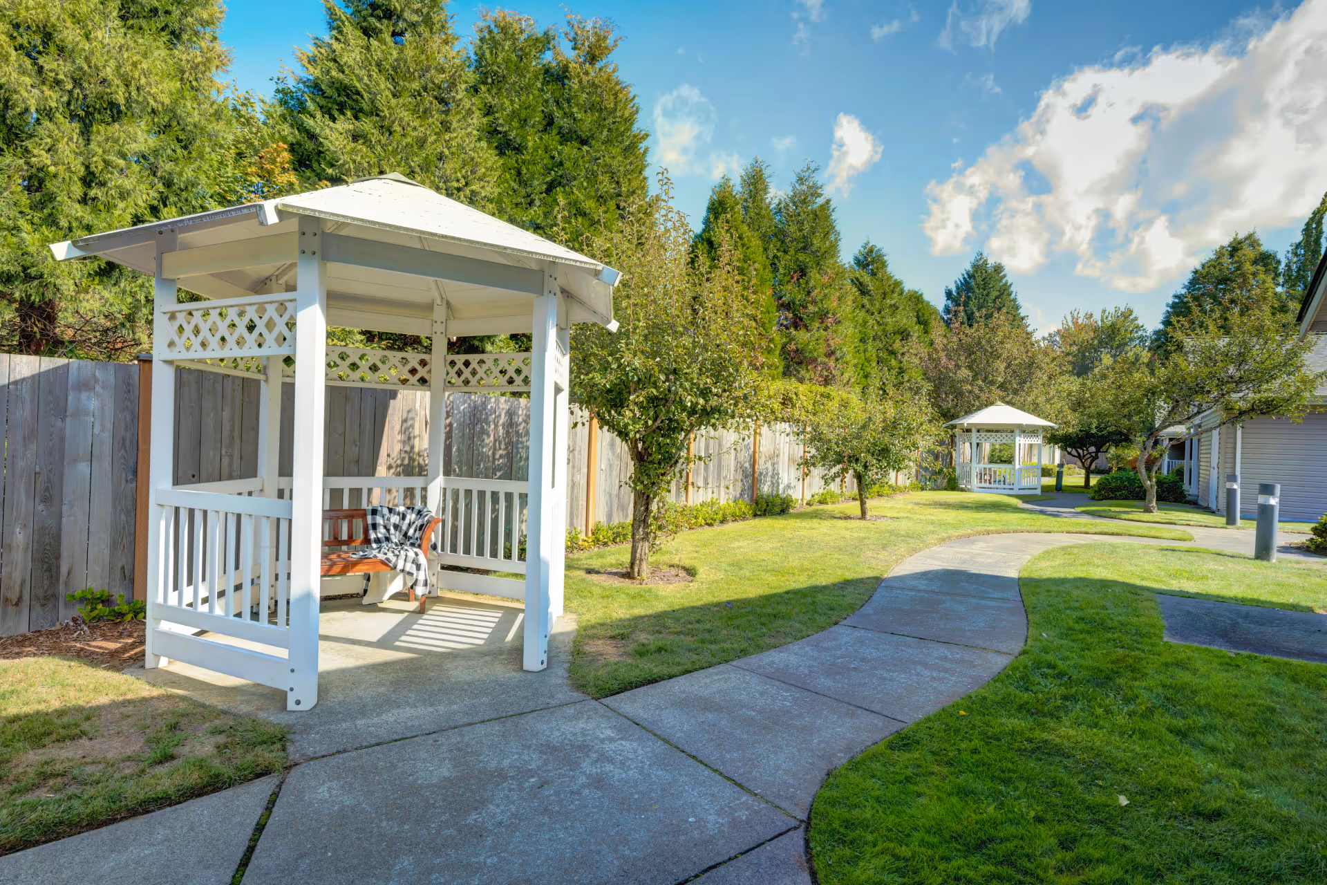 Paved walkway through a landscaped courtyard with white gazebos, benches, and trees under a blue sky.