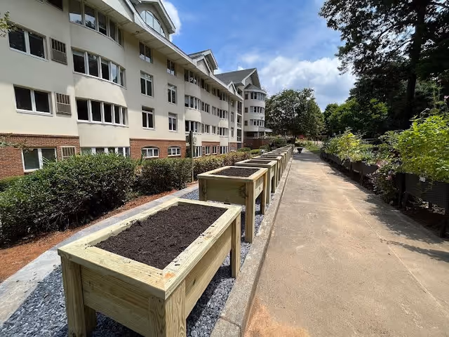 Outdoor walkway beside a multi-story residential building with a row of empty wooden planter boxes filled with soil on one side and green plants on the other side under a partly cloudy sky.