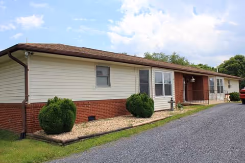Single-story senior living building with beige siding, brick accents, shrubs, and a gravel driveway.