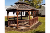 A wooden gazebo with a shingled roof situated on a grassy area outside, surrounded by a white fence and buildings in the background under a clear sky.