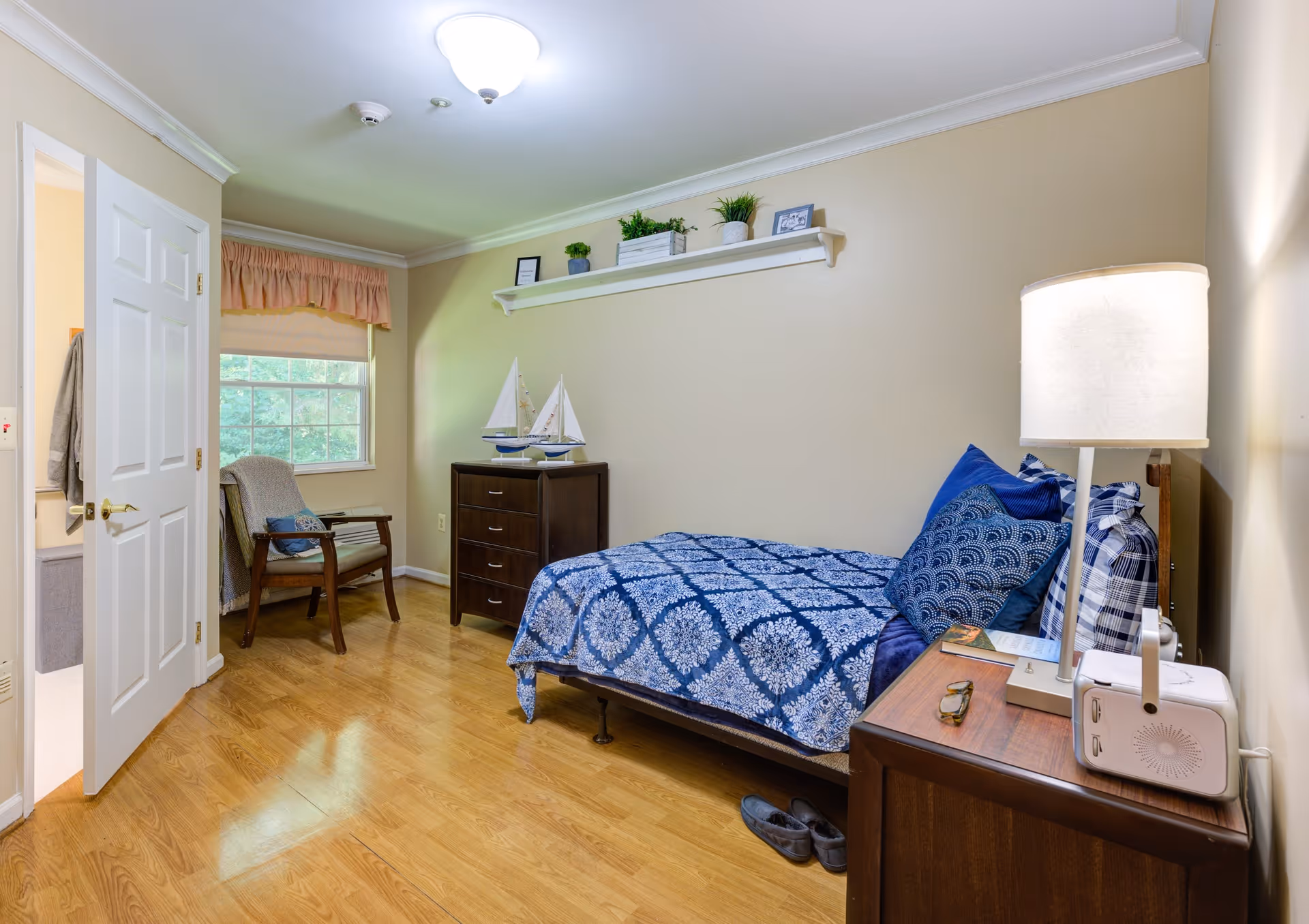 A cozy bedroom with a single bed covered in a blue patterned quilt and multiple pillows. Next to the bed is a wooden nightstand with a lamp, a small radio, and a pair of glasses. Across from the bed is a dark wooden dresser with two decorative sailboat models on top. A wooden chair with a cushion is placed near a window with a pink valance. The room has light beige walls, wooden flooring, and a ceiling light fixture. An open door leads to a bathroom.