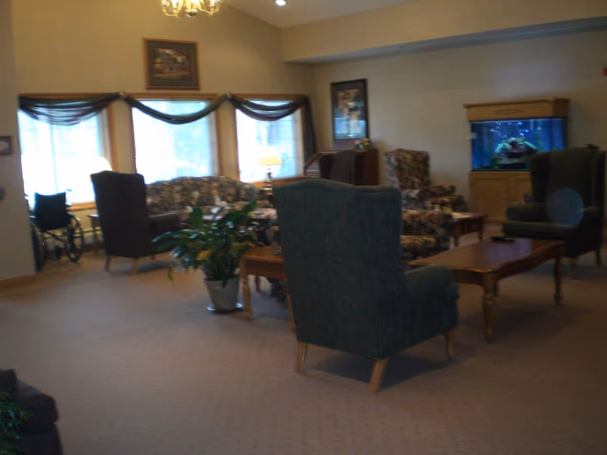 A cozy living room area with several upholstered armchairs and a floral-patterned sofa arranged around wooden coffee tables. There is a large fish tank on a wooden cabinet against the wall, a potted plant on the floor, and windows with sheer curtains letting in natural light. The room has beige walls and carpeted flooring.