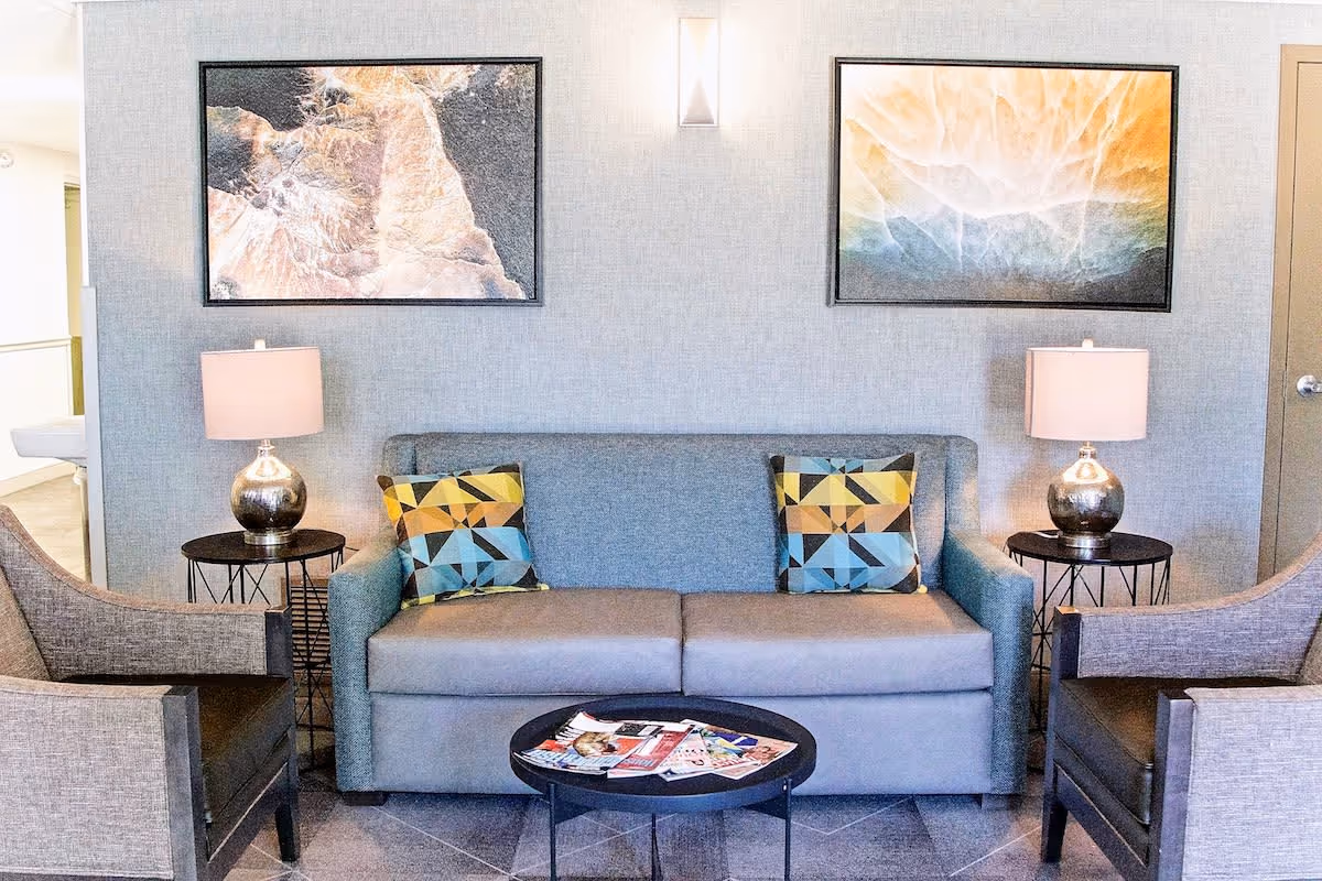 A modern living room area with a gray sofa adorned with two geometric patterned pillows, flanked by two matching armchairs. Each side of the sofa has a round side table with a metallic lamp. Two abstract paintings hang on the light gray wall above the sofa, and a small round coffee table with magazines is placed in front of the sofa.