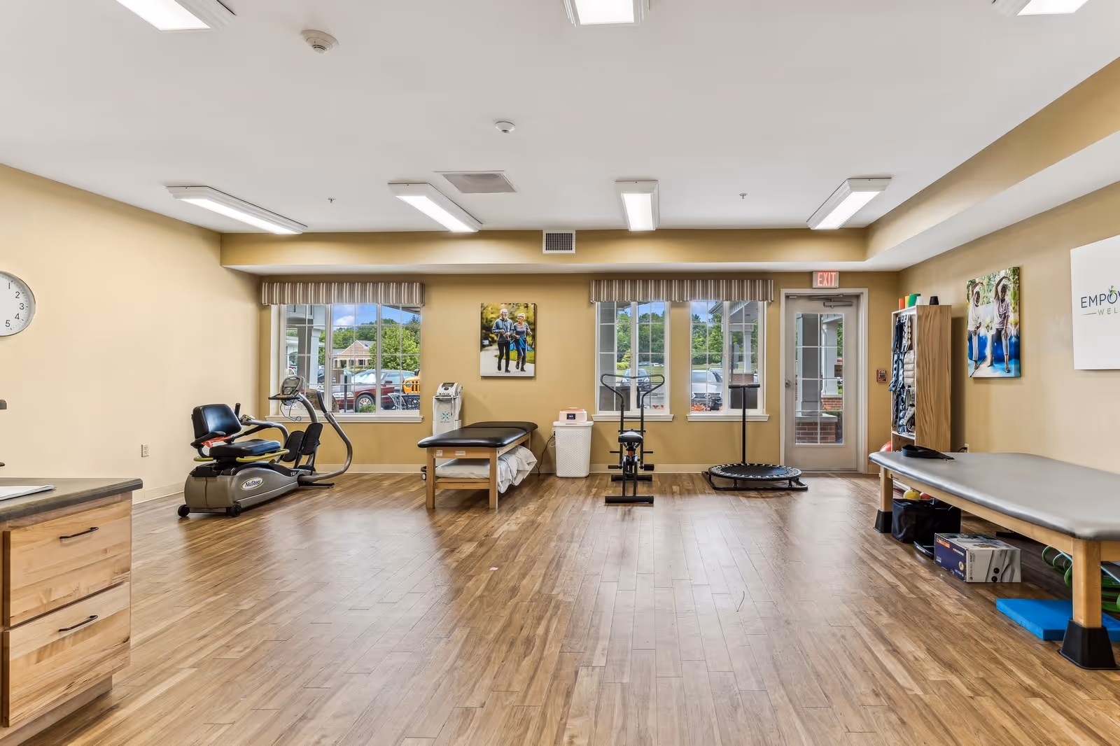 A spacious room with wooden flooring and beige walls, featuring exercise equipment including a recumbent bike, stationary bike, and a small trampoline. There are two treatment tables, windows with striped valances, and framed pictures on the walls. The room is well-lit with ceiling lights and has a door leading outside.
