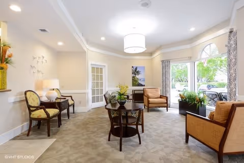 Bright lounge with armchairs, side tables, plants, and large windows overlooking greenery.