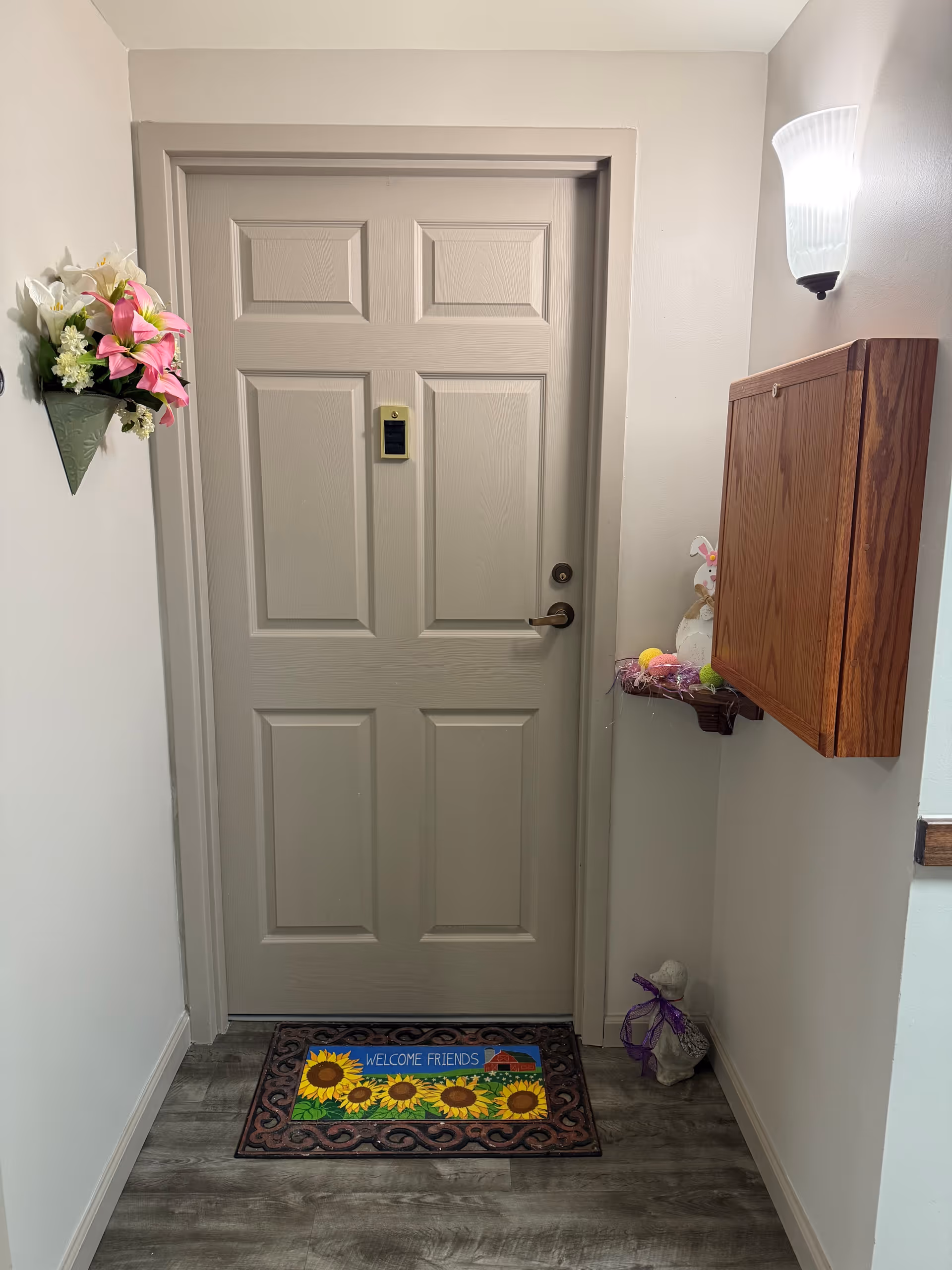 A beige door with a silver handle in a hallway. A colorful doormat with sunflowers and the text 'WELCOME FRIENDS' is placed in front of the door. On the left wall, there is a decorative wall-mounted vase with pink and white flowers. On the right wall, there is a wooden cabinet above a small shelf decorated with Easter-themed items including a bunny figure and colorful eggs. A wall-mounted light fixture is above the cabinet, illuminating the area.
