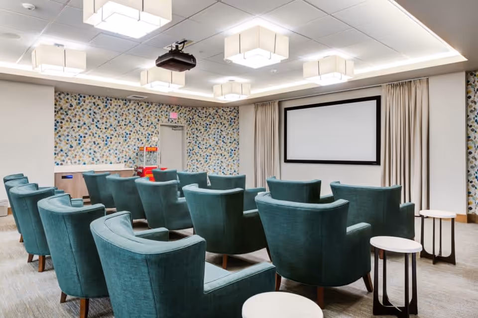 A small theater room with multiple teal upholstered armchairs arranged in rows facing a large blank projection screen. The room has a patterned accent wall with blue, beige, and gray dots, ceiling-mounted projector, modern square ceiling lights, and small round side tables next to some chairs.