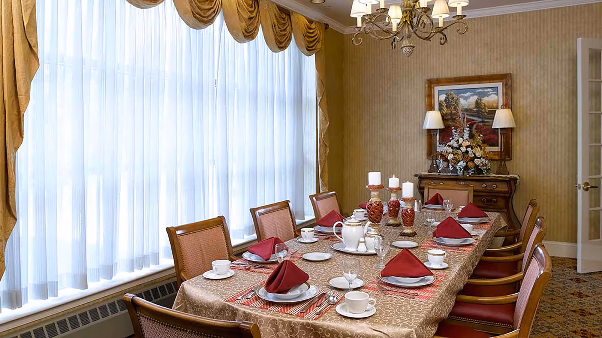 A formal dining room with a long table set for eight people. The table is covered with a patterned tablecloth and set with white plates, cups, silverware, and red folded napkins. There are decorative candles and a tea set in the center of the table. The room has large windows with sheer white curtains and gold drapes, a chandelier overhead, and a sideboard with a floral arrangement and a framed landscape painting on the wall.