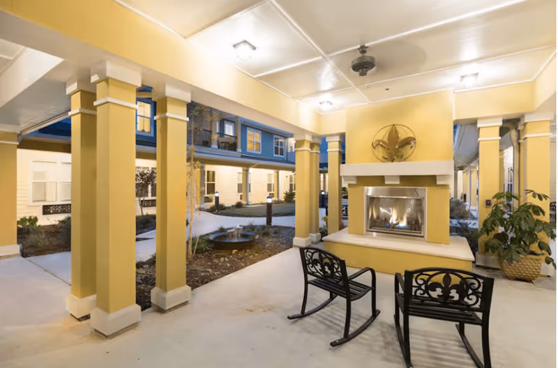 Covered courtyard with columns, a lit outdoor fireplace and two decorative metal rocking chairs in front of the senior living building.
