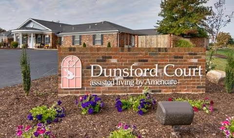 Brick sign for Dunsford Court assisted living by Americare surrounded by mulch and purple flowers, with a building and parking lot visible in the background under a partly cloudy sky.