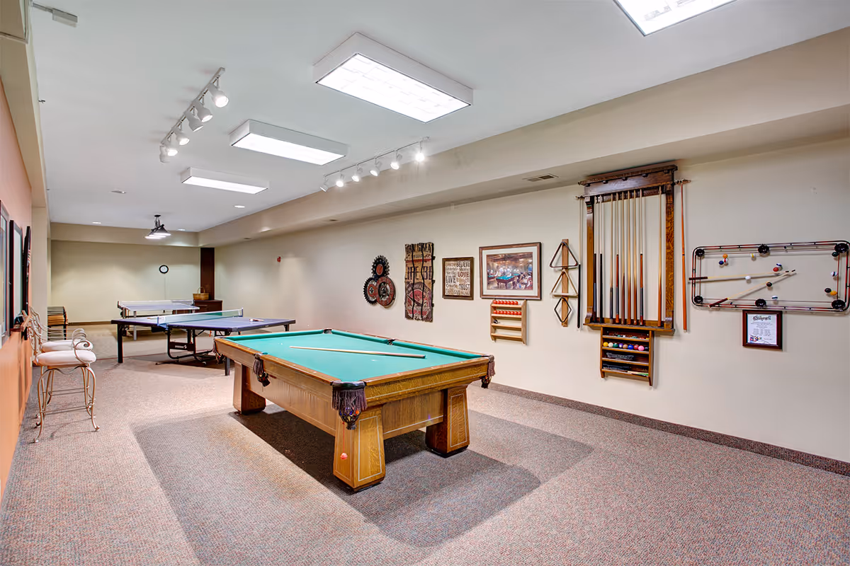 Recreation room with a pool table in the foreground, ping-pong tables in the background, and cue racks and wall decor along the right wall.