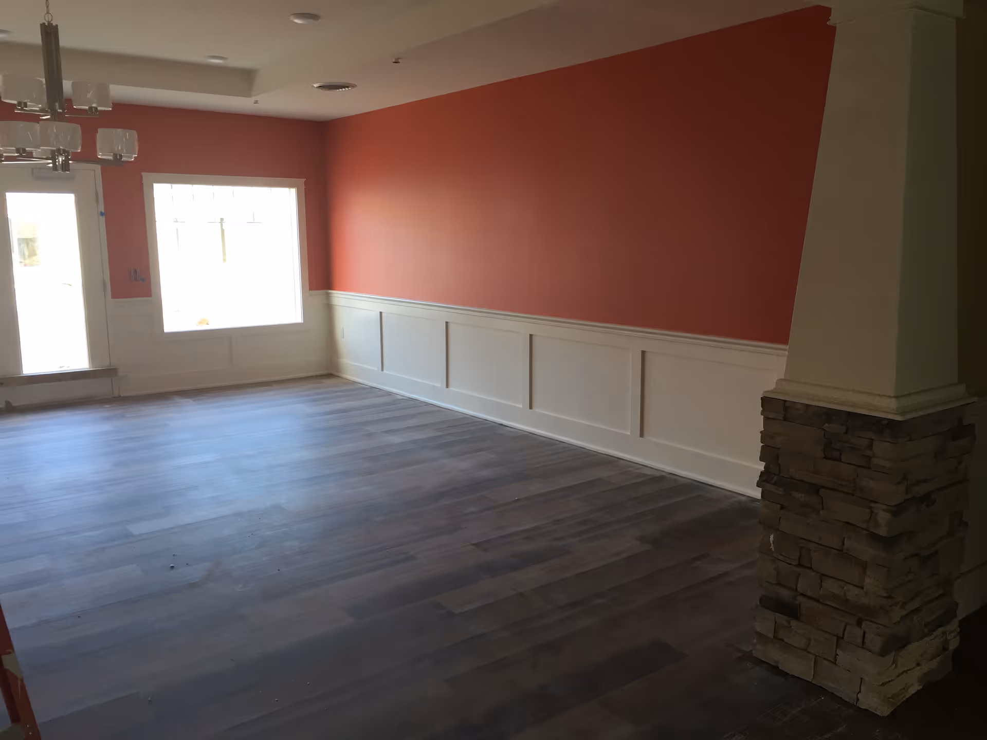 Empty interior room with dark wood flooring, coral-colored walls with white wainscoting, a large window, a glass door, a modern chandelier, and a stone pillar on the right side.