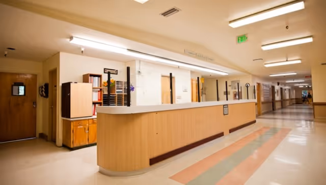 A wide hallway in a healthcare facility with a long wooden reception desk on the left side. The hallway is well-lit with fluorescent ceiling lights and has a patterned floor with beige, green, and orange tiles. Doors and cabinets are visible along the walls, and an exit sign is illuminated in green above one door.