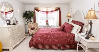 A cozy bedroom featuring a large bed with red bedding and multiple pillows, a white wooden headboard, two bedside tables each with a lamp, a white dresser with a round mirror, a patterned armchair, a potted plant, and a window with sheer and red curtains allowing natural light to fill the room.