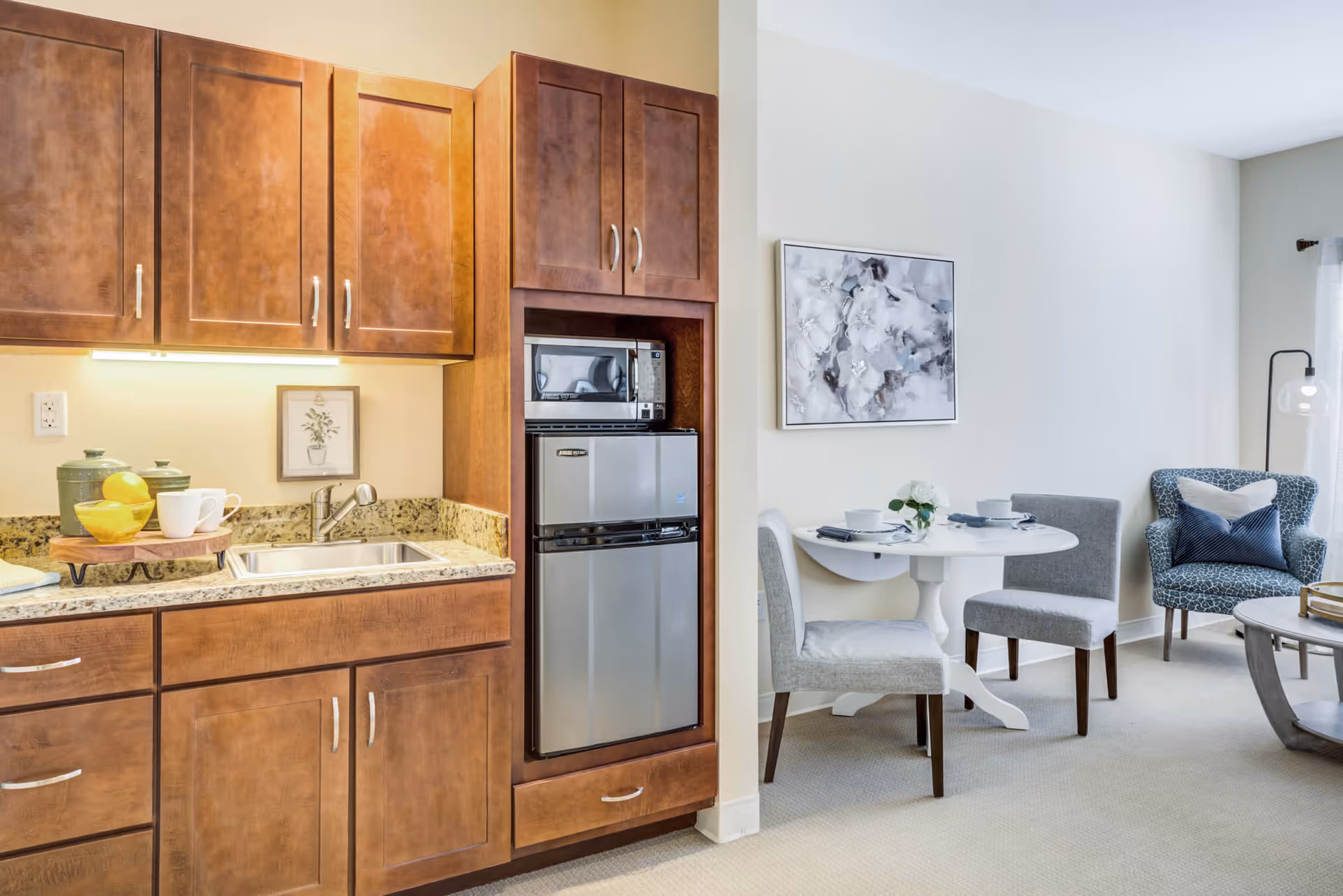 A small kitchen area with wooden cabinets, a granite countertop, a sink, a microwave, and a mini refrigerator. Adjacent to the kitchen is a dining area with a round white table set for two, two gray upholstered chairs, and a blue patterned armchair. A floral painting hangs on the wall above the dining table.