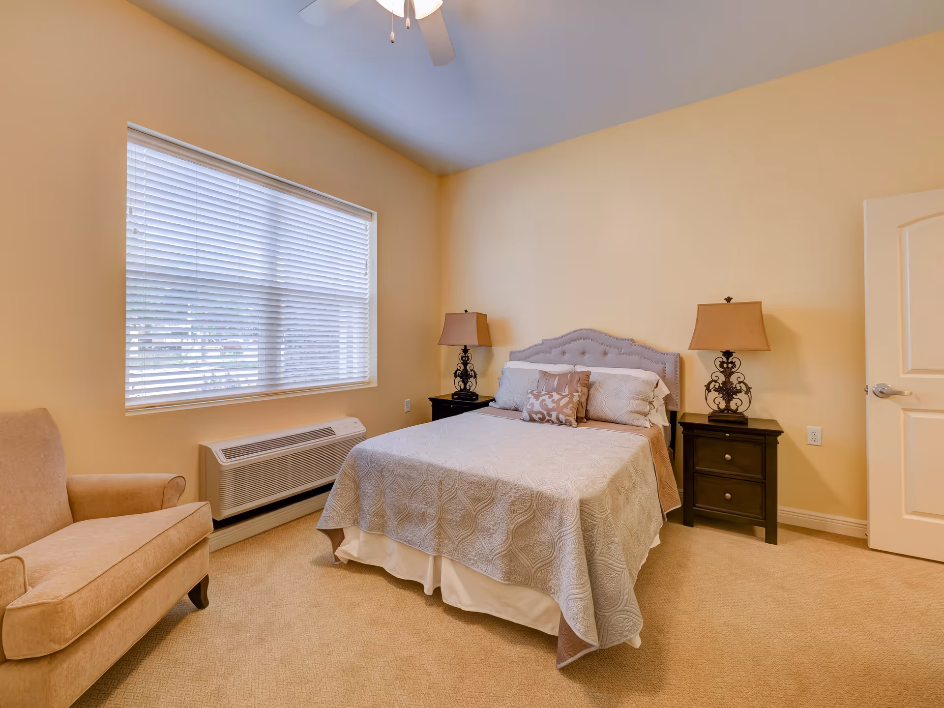 A cozy bedroom with a neatly made bed featuring a light gray quilt and decorative pillows. There are two dark wooden nightstands on either side of the bed, each with an ornate lamp. A beige armchair is positioned near a large window with white blinds, and the walls are painted a soft yellow color. The room has beige carpeting and a white door with a silver handle.