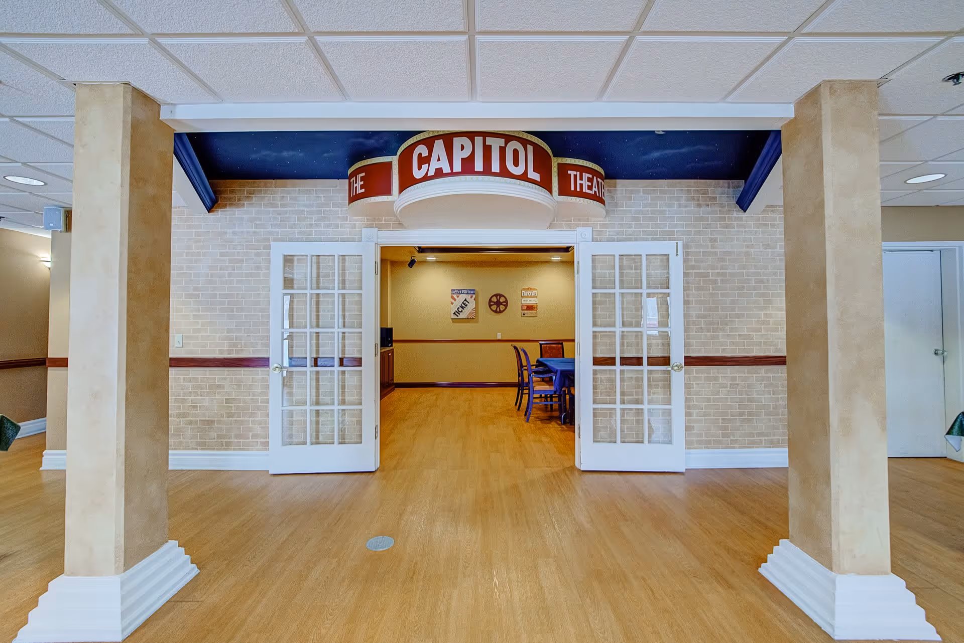 Interior view of a room entrance labeled 'The Capitol Theatre' with double glass doors open, showing a room with wooden flooring, beige brick walls, and a table with chairs inside. Two beige columns frame the entrance.