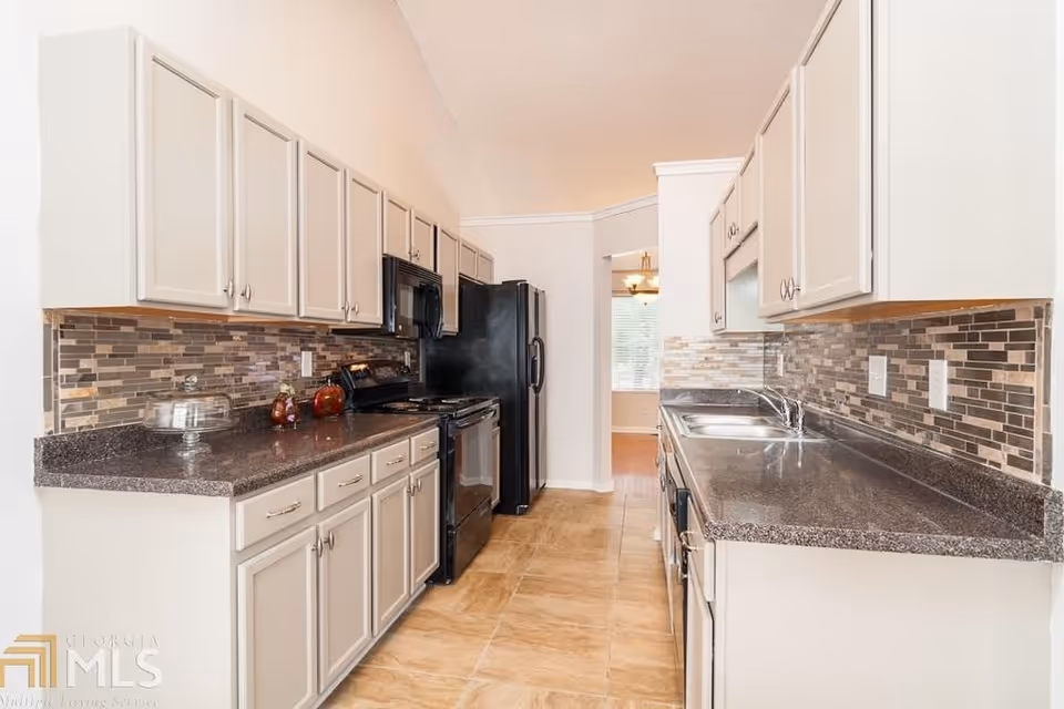 A modern kitchen with beige cabinets and dark countertops on both sides. The kitchen features a black refrigerator, black microwave, black stove, and a stainless steel sink. The backsplash is made of rectangular tiles in shades of brown and beige. The floor is tiled in a light brown color, and there is a doorway leading to a dining area with a window and a hanging light fixture.