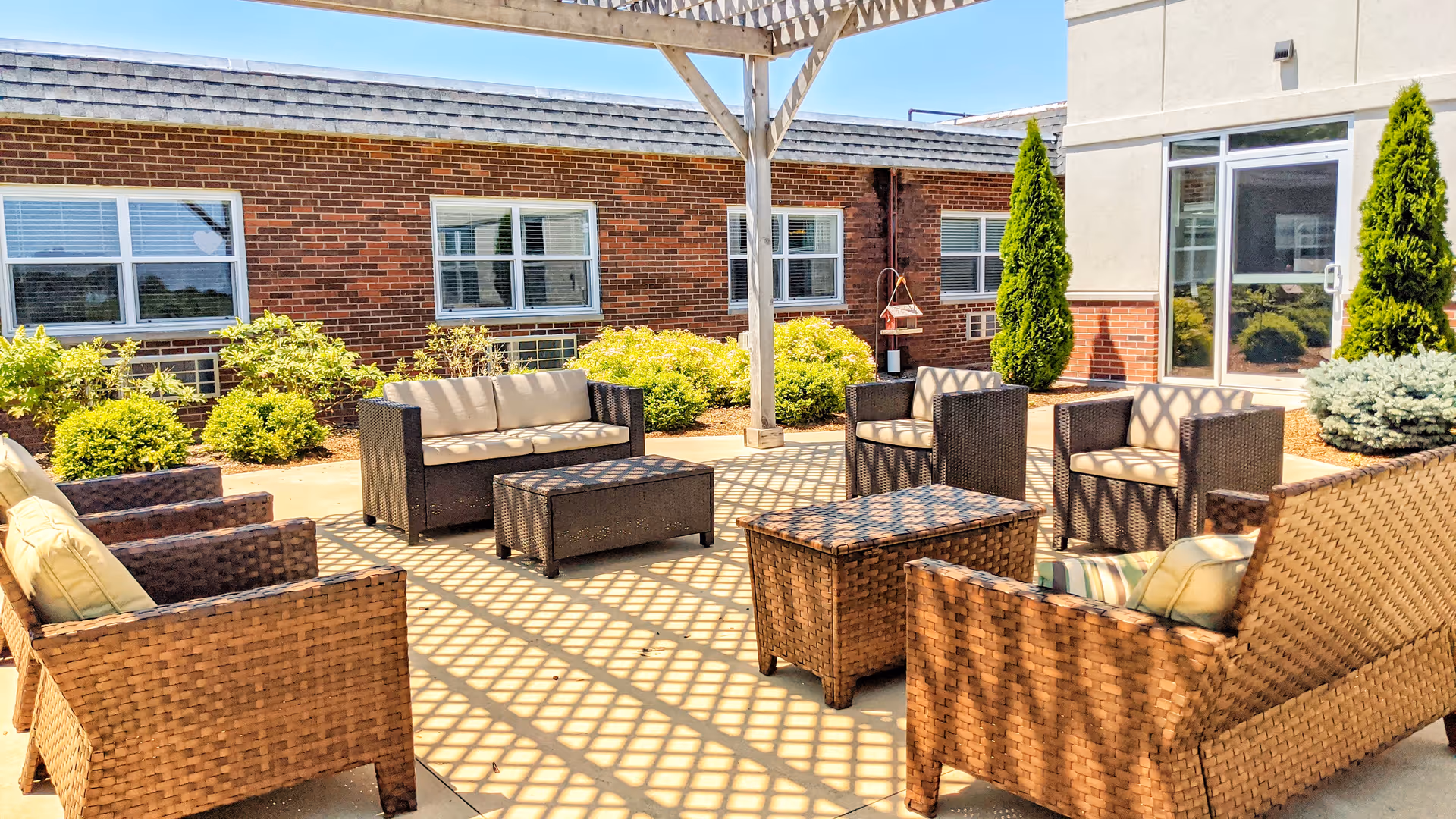 Outdoor patio area with wicker furniture including sofas, armchairs, and tables arranged under a wooden pergola casting a lattice shadow. The patio is surrounded by bushes and small trees, with a brick building and glass door in the background under a clear blue sky.