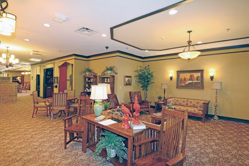 Well-lit communal lounge with wooden tables and chairs, sofas, lamps, and decorative plants.