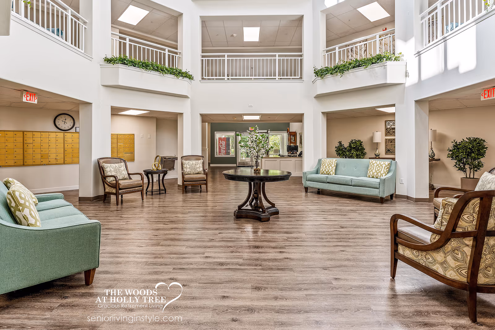 Spacious and well-lit senior living facility lobby with wooden flooring, two green sofas with patterned cushions, four armchairs, a round wooden table with a flower arrangement in the center, and mailboxes on the left wall. The area features white walls, a high ceiling with a second-floor balcony, and exit signs above doorways.