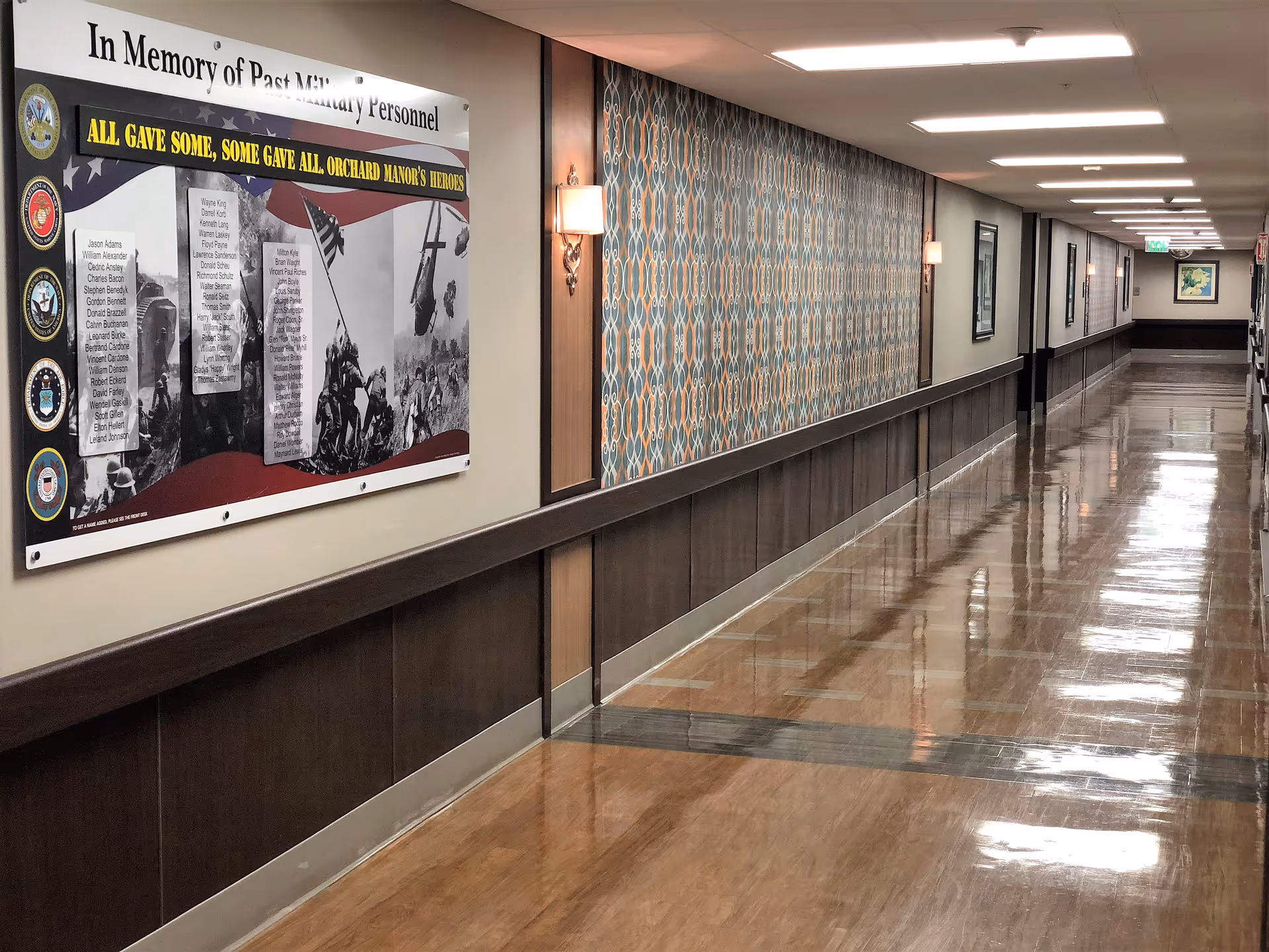 A long, polished hallway in a senior living facility with wood paneling on the lower half of the walls and patterned wallpaper above. The hallway is well-lit with ceiling lights and wall sconces. On the left wall, there is a large memorial poster honoring past military personnel with names and military branch insignias. Several framed pictures are visible further down the hallway.