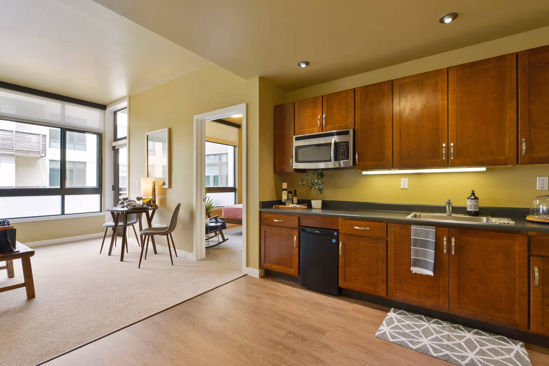 Interior view of a senior living facility apartment showing a kitchen area with wooden cabinets, a microwave, a small refrigerator, and a sink with a towel hanging. Adjacent to the kitchen is a dining area with a small round table and two chairs near large windows. A doorway leads to a bedroom with a bed and a chair visible.