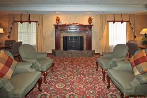 A cozy living room with a patterned red carpet, two green upholstered armchairs with plaid cushions facing each other, a wooden fireplace mantel decorated with autumn-themed floral arrangements, and windows with beige curtains on either side.