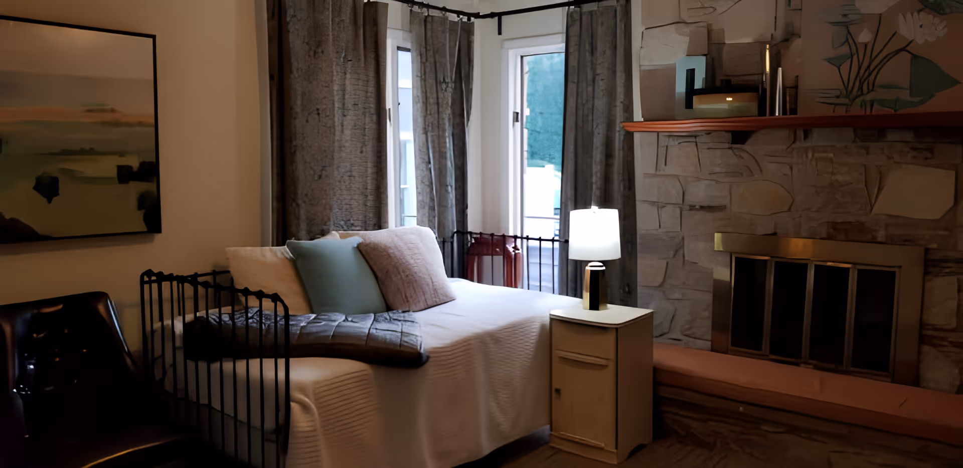 A cozy living room area with a white daybed adorned with pillows and a blanket, positioned near two tall windows with gray curtains. A small white side table with a lit lamp sits next to the daybed. The room features a stone fireplace with a wooden mantel decorated with various items, and a cushioned bench in front of the fireplace. A dark chair is partially visible on the left side, and a framed abstract painting hangs on the wall.