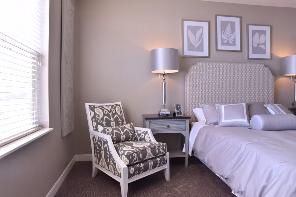 A cozy bedroom with a patterned armchair next to a window with blinds. The bed has a light-colored headboard and is adorned with multiple pillows and a bolster. There are two bedside tables, each with a lamp and small framed photos. Three framed botanical prints hang on the wall above the bed.