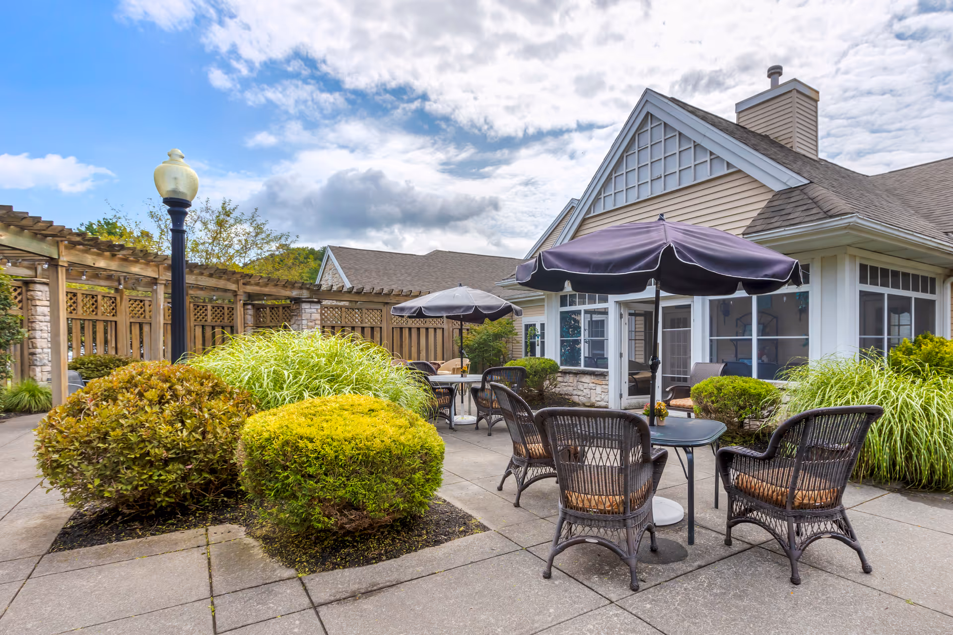 Outdoor courtyard patio with wicker tables and chairs under umbrellas, landscaped shrubs, and the facility building in the background.