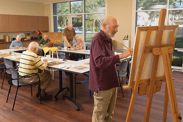 Several elderly residents paint and draw at tables in a bright activity room while a man works at an easel in the foreground.