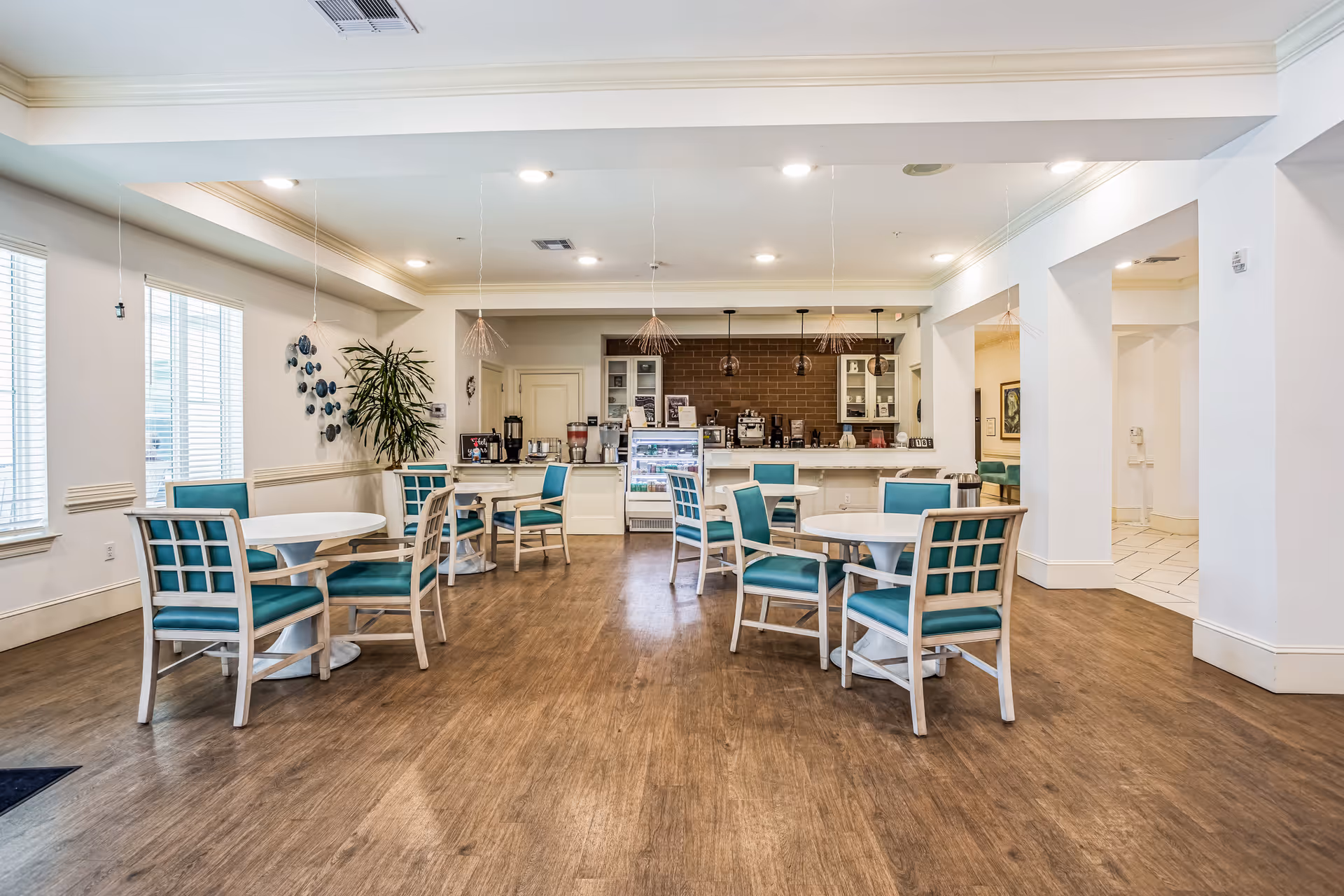 A bright and spacious dining area with several round white tables and teal cushioned chairs. The room has large windows with blinds on the left side, wooden flooring, and white walls. At the back, there is a counter area with coffee machines, a display fridge, and hanging pendant lights. A potted plant and wall decor are visible on the left wall.