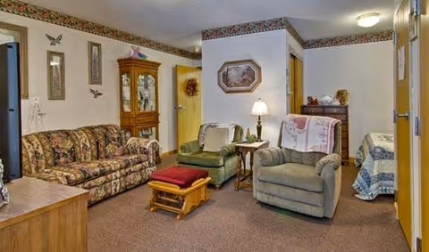 A cozy living room area in a senior living facility with a floral patterned sofa, two armchairs with white doilies on the backrests, a wooden rocking footstool, a side table with a lamp, a wooden cabinet with glass doors, and framed wall decorations including butterflies and a wreath.