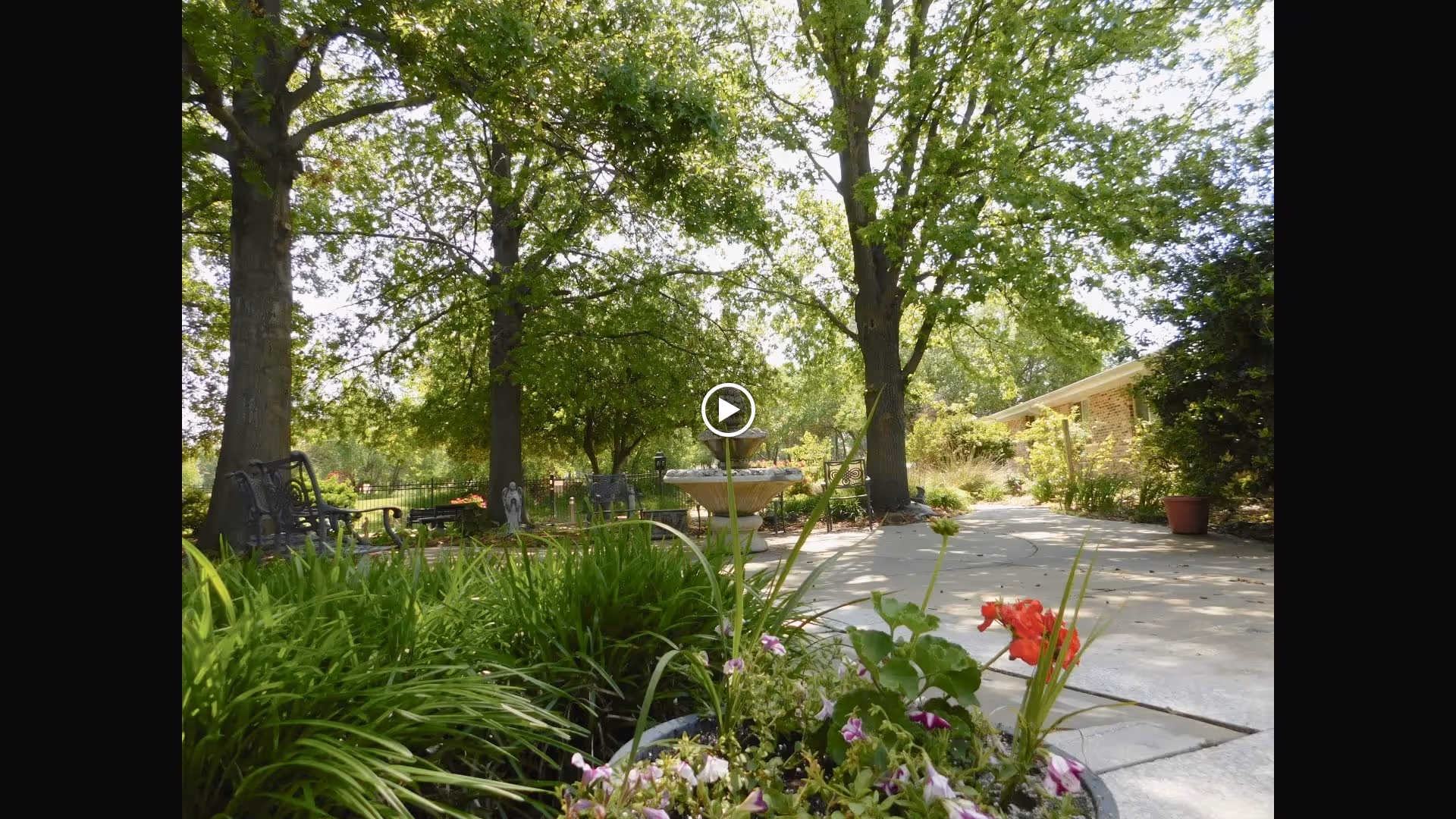 A peaceful outdoor garden area with lush green plants and flowers in the foreground, large trees providing shade, a stone birdbath in the center, metal benches on the left side, and a building partially visible on the right side under a bright sky.
