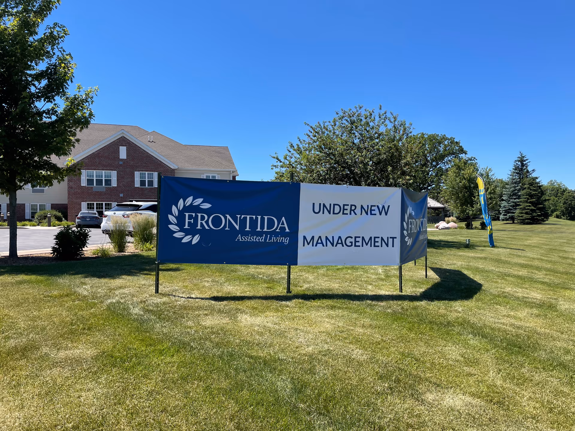 A large blue and white banner on a grassy lawn in front of a brick and beige building. The banner reads 'FRONTIDA Assisted Living UNDER NEW MANAGEMENT'. There are trees, parked cars, and a clear blue sky in the background.