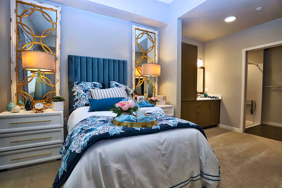 A neatly made bed with a blue upholstered headboard and floral bedding in a senior living bedroom. Two decorative mirrors with gold geometric frames and table lamps are on either side of the bed on white nightstands. A tray with flowers and a cup is placed on the bed. To the right, there is a vanity area with a sink and a mirror, and an open doorway leading to a walk-in closet and bathroom.