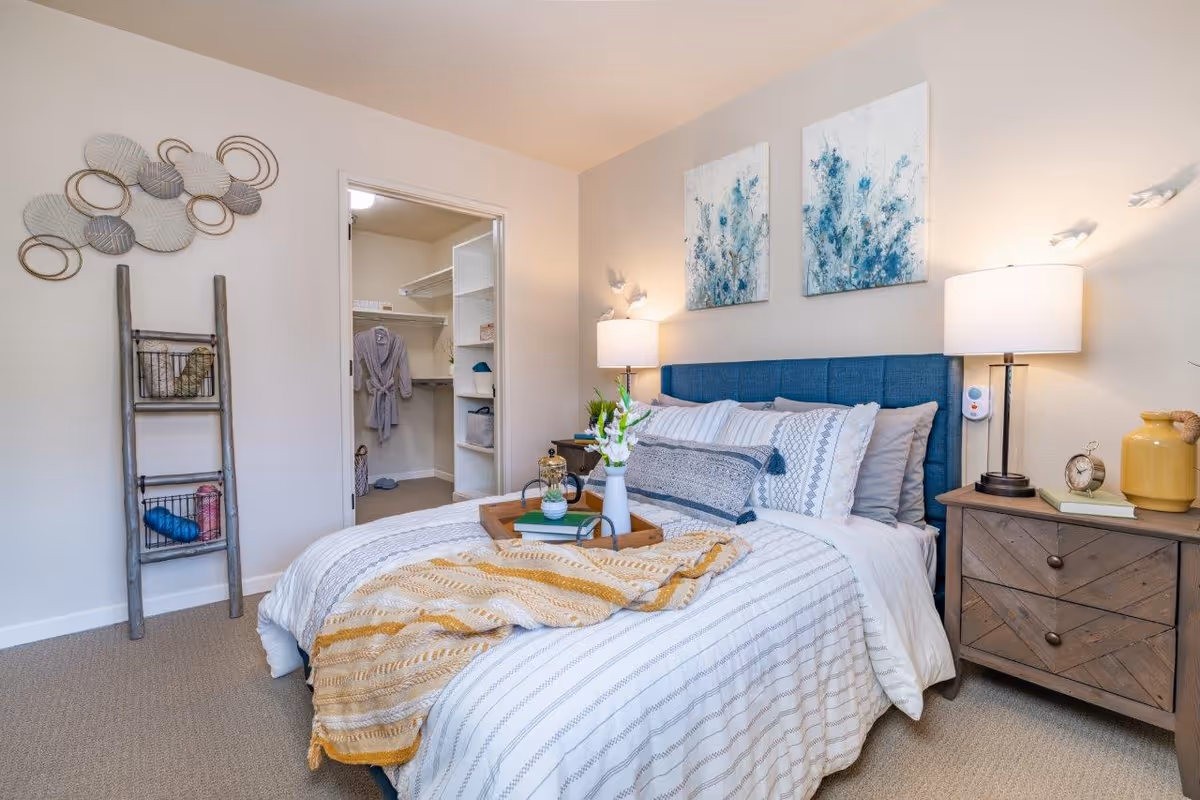 Well-lit bedroom with a blue upholstered headboard, a neatly made bed with patterned linens, bedside tables and lamps, wall art, a decorative ladder, and an open walk-in closet.