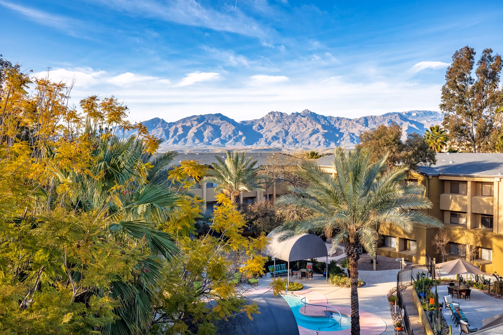 Outdoor courtyard with palm trees, a small pool, and seating beside a multi-story assisted living building with mountains in the background.