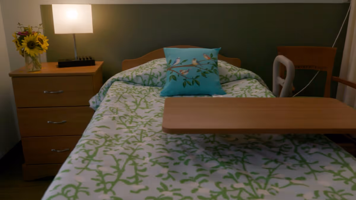 A single bed in a nursing facility room with a green and white patterned bedspread and a decorative pillow featuring birds on branches. Next to the bed is a wooden nightstand with a lamp and a vase of sunflowers. A wooden overbed table is positioned over the bed.