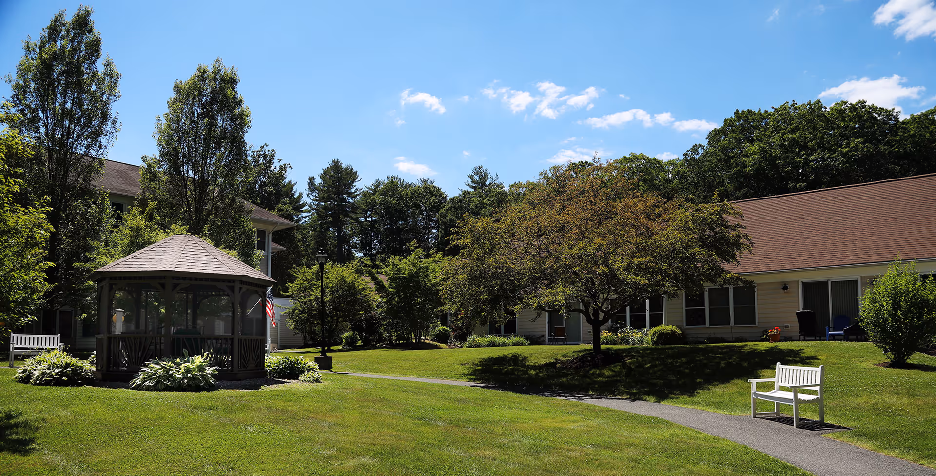 A sunny outdoor area at Rockridge Retirement Community featuring a green lawn, a gazebo, trees, benches, and buildings with brown roofs under a blue sky with some clouds.