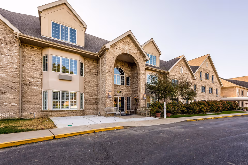 Exterior view of a large brick assisted living facility with multiple windows, a covered entrance with an archway, benches near the entrance, and some landscaping including bushes and small trees.