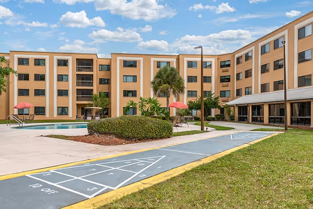 Outdoor courtyard area of a senior living facility with a shuffleboard court in the foreground, a swimming pool with red umbrellas and lounge chairs, surrounded by a multi-story beige building under a partly cloudy blue sky.