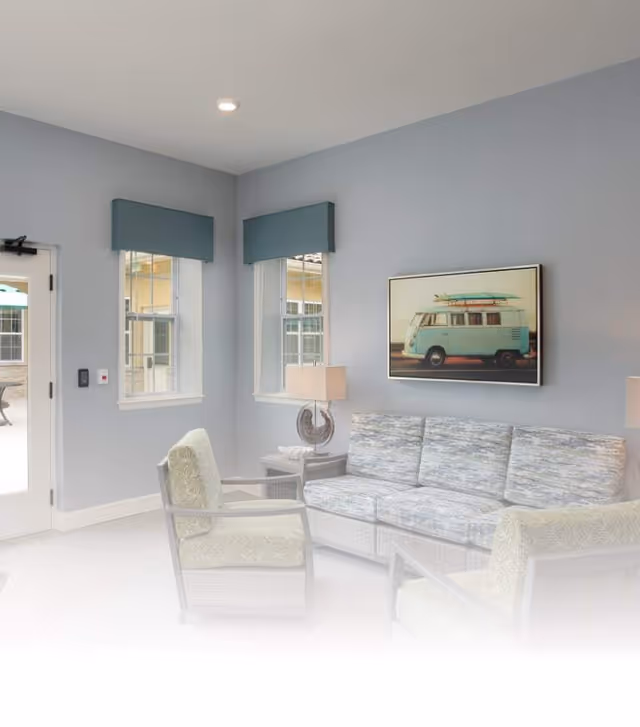 A cozy living room area with a light blue-gray wall, two windows with blue valances, a framed picture of a vintage van with surfboards on top, a cushioned sofa with a patterned fabric, two cushioned armchairs, and two table lamps on side tables. A glass door leads outside to a patio area.