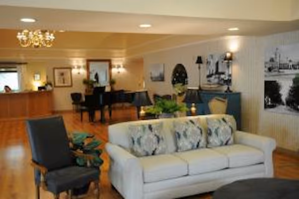 A spacious and well-lit living room area with a white sofa adorned with patterned cushions, a dark upholstered armchair, a grand piano in the background, wooden flooring, and various decorative elements including framed pictures, plants, and lamps.