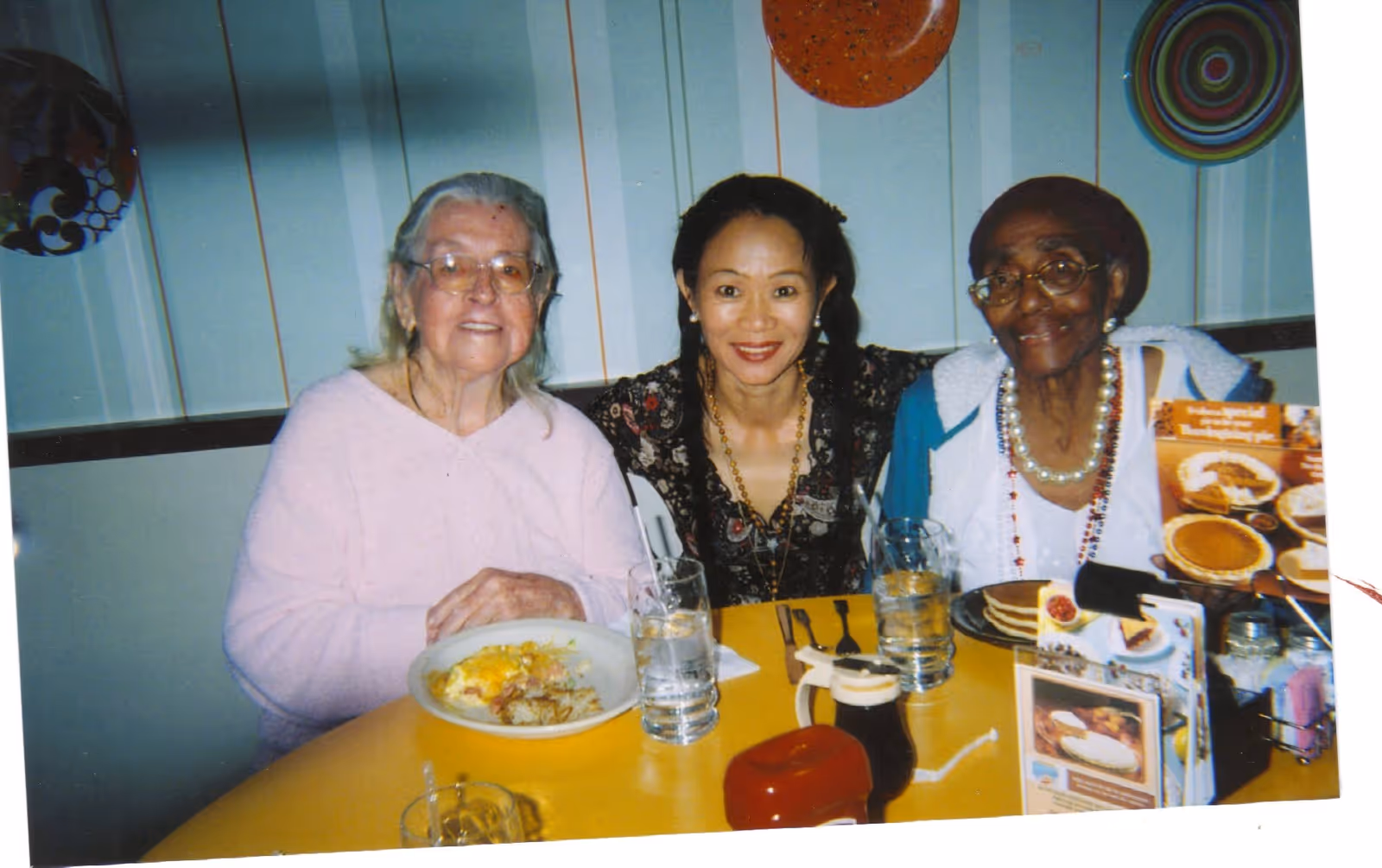 Three women sitting at a yellow table in a dining area. Two elderly women are seated on either side of a younger woman in the middle. The table has plates of food, glasses of water, a syrup dispenser, and menus. The background features a wall with decorative plates hanging.