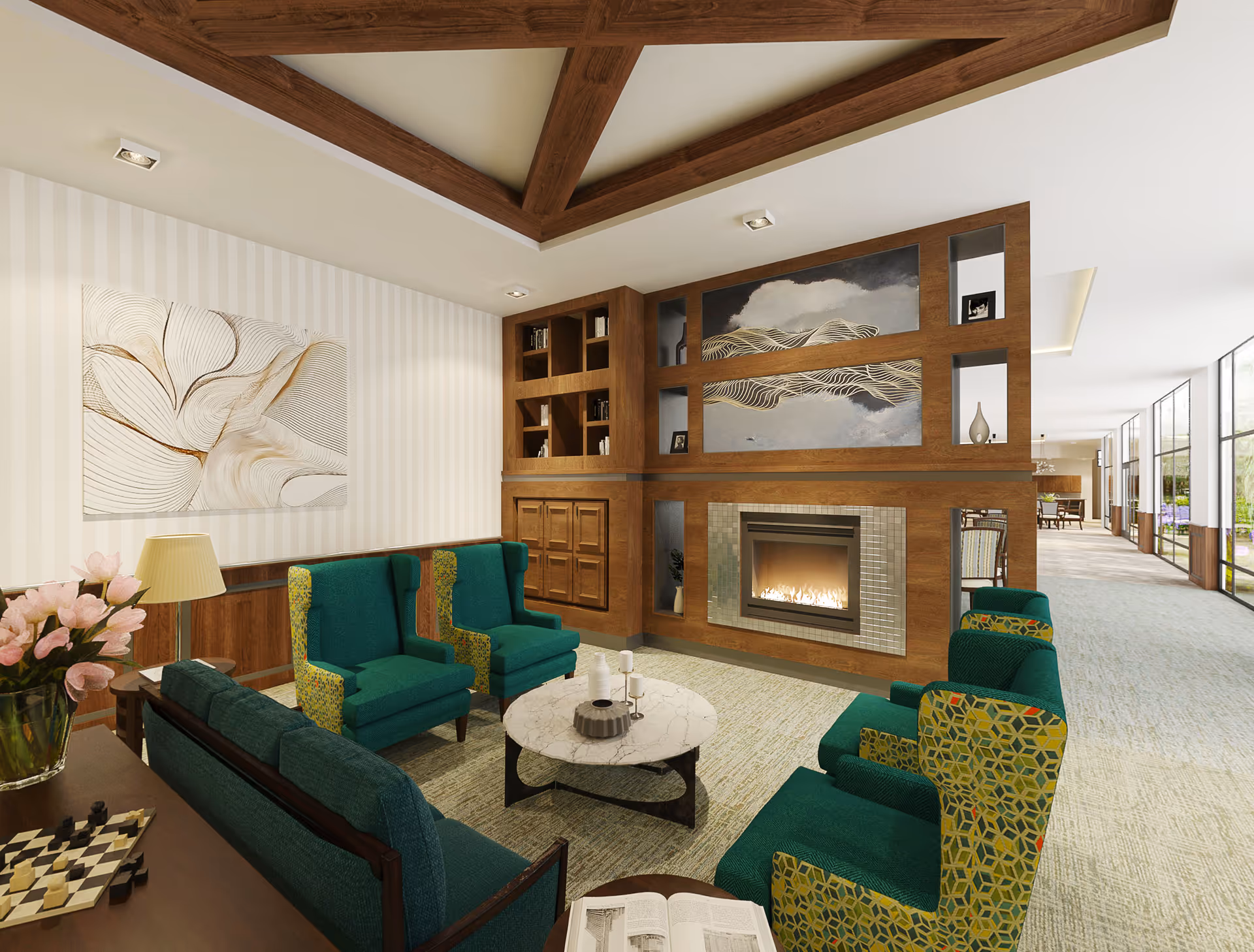 A cozy living room area with green upholstered armchairs and a matching sofa arranged around a round marble coffee table. The room features a wooden built-in shelving unit with a fireplace in the center, decorated with artwork and small decorative items. There is a side table with a lamp and a vase of pink flowers, and a chessboard is visible on a nearby surface. Large windows line the hallway visible in the background, allowing natural light to fill the space.
