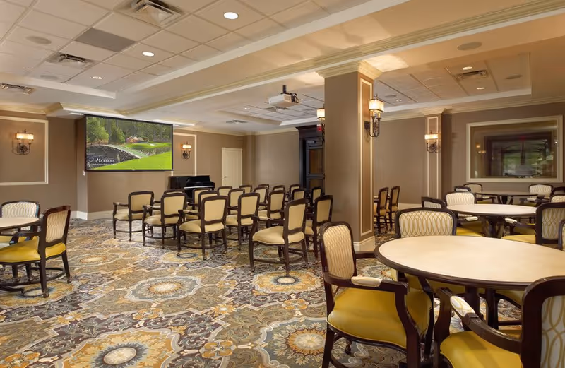 A spacious meeting or activity room with multiple round tables and chairs arranged in rows facing a large screen on the wall. The room features patterned carpet flooring, beige walls with decorative molding, wall sconces for lighting, and a piano in the corner.