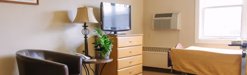 A cozy senior living room featuring a brown leather armchair, a round side table with a lamp and a potted plant, a wooden dresser with a flat-screen TV on top, a bed with a yellow bedspread, a wall-mounted air conditioning unit, and a window letting in natural light.