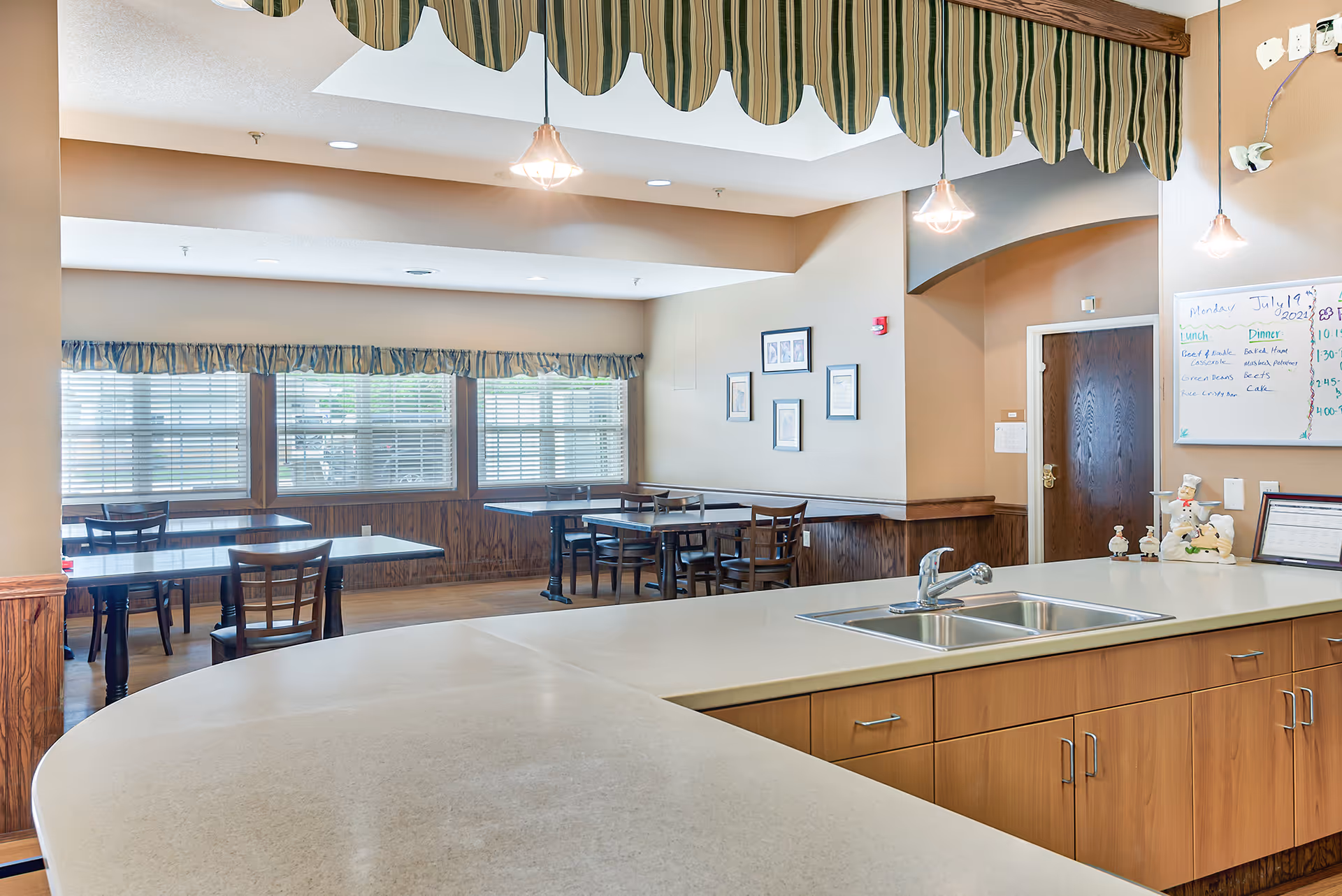 Interior view of a senior living facility dining area with several tables and chairs near windows with blinds and valances. In the foreground, there is a large countertop with a double sink and wooden cabinets underneath. The walls are beige with framed pictures and a whiteboard displaying a menu and schedule. Pendant lights hang from the ceiling.